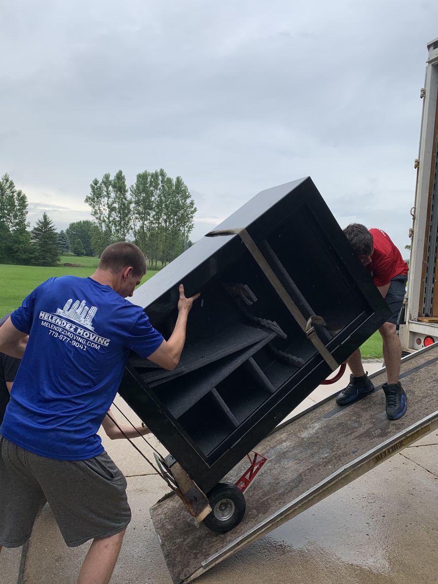 Melendez Moving crew loading a wrapped, strapped cabinet up a truck ramp with a hand truck